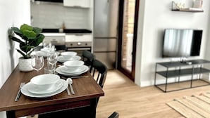 A modern dining area with a wooden table set for four, a kitchen in the background, and a TV on a stand.