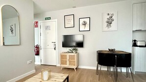 Un salon moderne avec une table basse en bois, une table à manger noire avec des chaises, un téléviseur et des reproductions botaniques encadrées accrochées au mur.