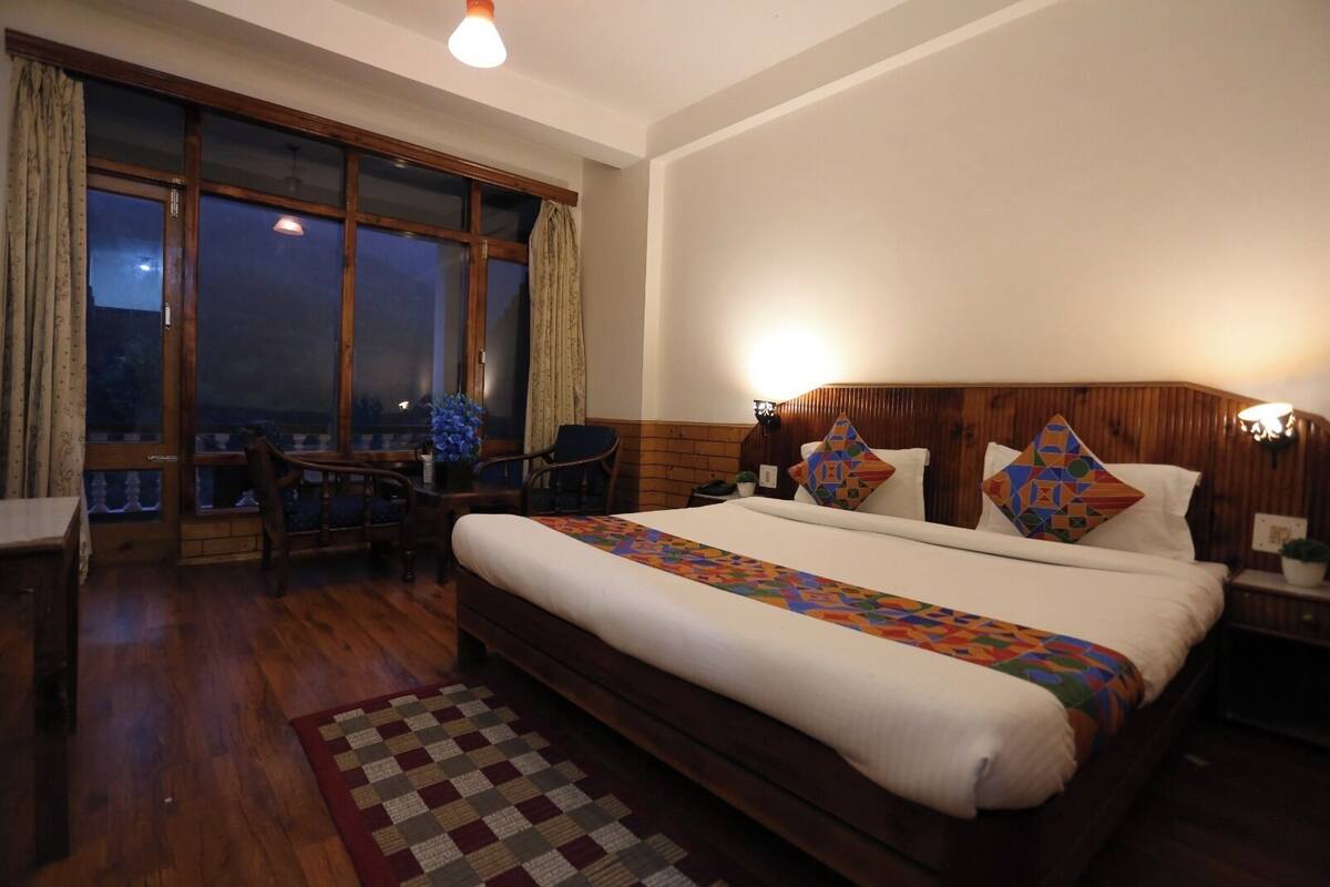 A hotel room with a large bed, wooden headboard, two bedside tables, a desk with a chair, a window with curtains, and a view of the outside.