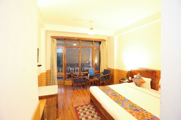 A hotel room with a large bed, a wooden floor, a balcony with a table and chairs, and a view of the outside.