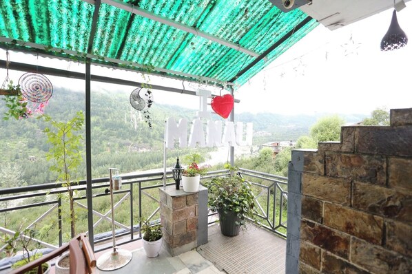 A balcony with a view of a mountainous landscape, a sign reading "MANAU", and potted plants.