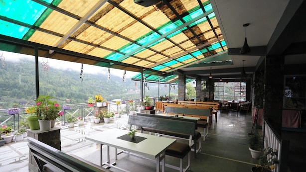 Un espacio al aire libre con mesas y bancos, rodeado de plantas en macetas y vistas a un paisaje montañoso.