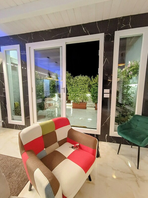 A modern living room with a checkered sofa, a green chair, and a glass door leading to an outdoor area with potted plants. - Blumoon  (Andria)