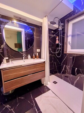 A modern bathroom with a marble vanity, a round mirror, a handheld showerhead, and a window with a frosted glass panel. - Blumoon  (Andria)