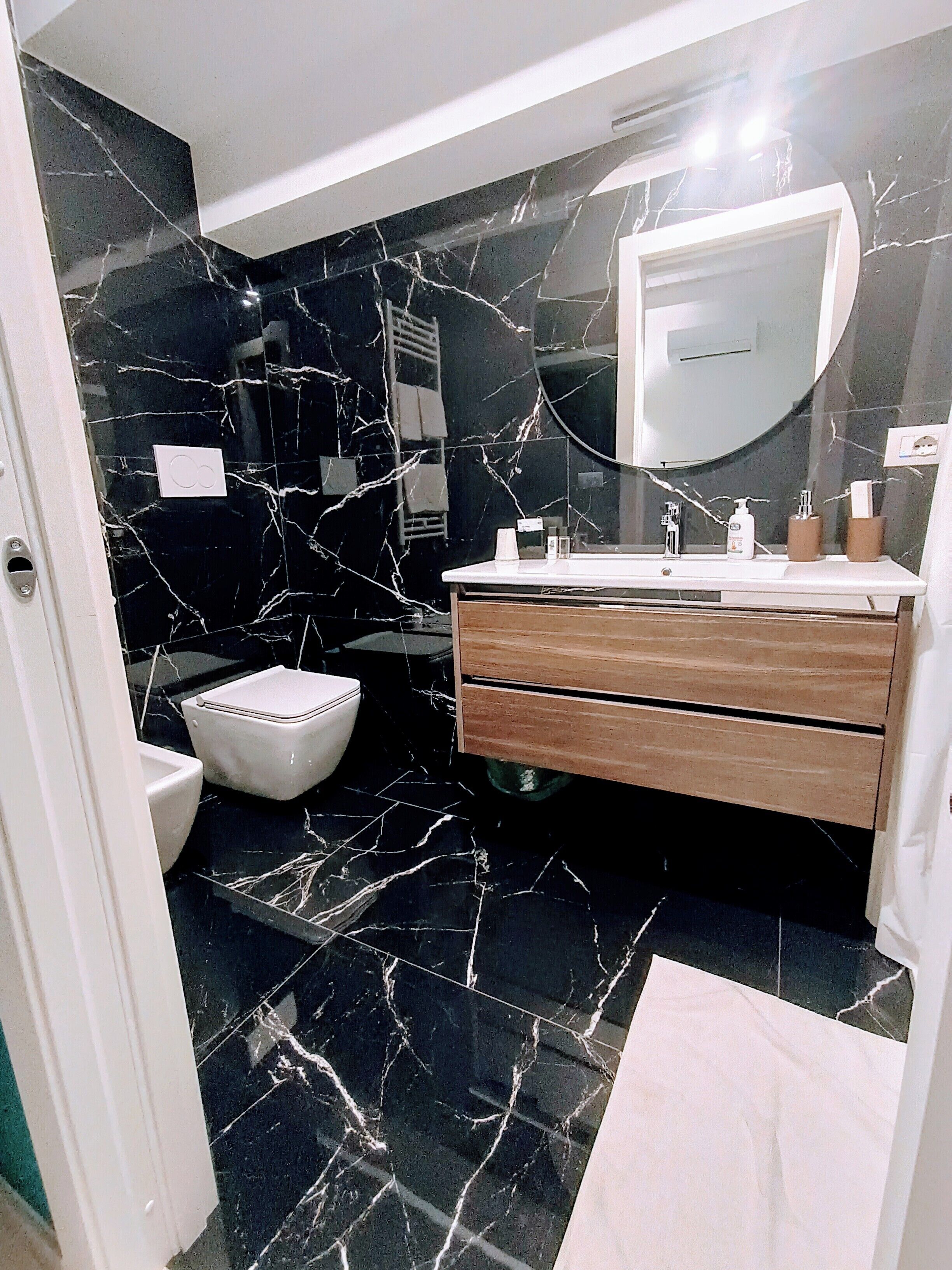 A modern bathroom with black marble walls, a white toilet, a wooden vanity with a mirror, and a white sink.