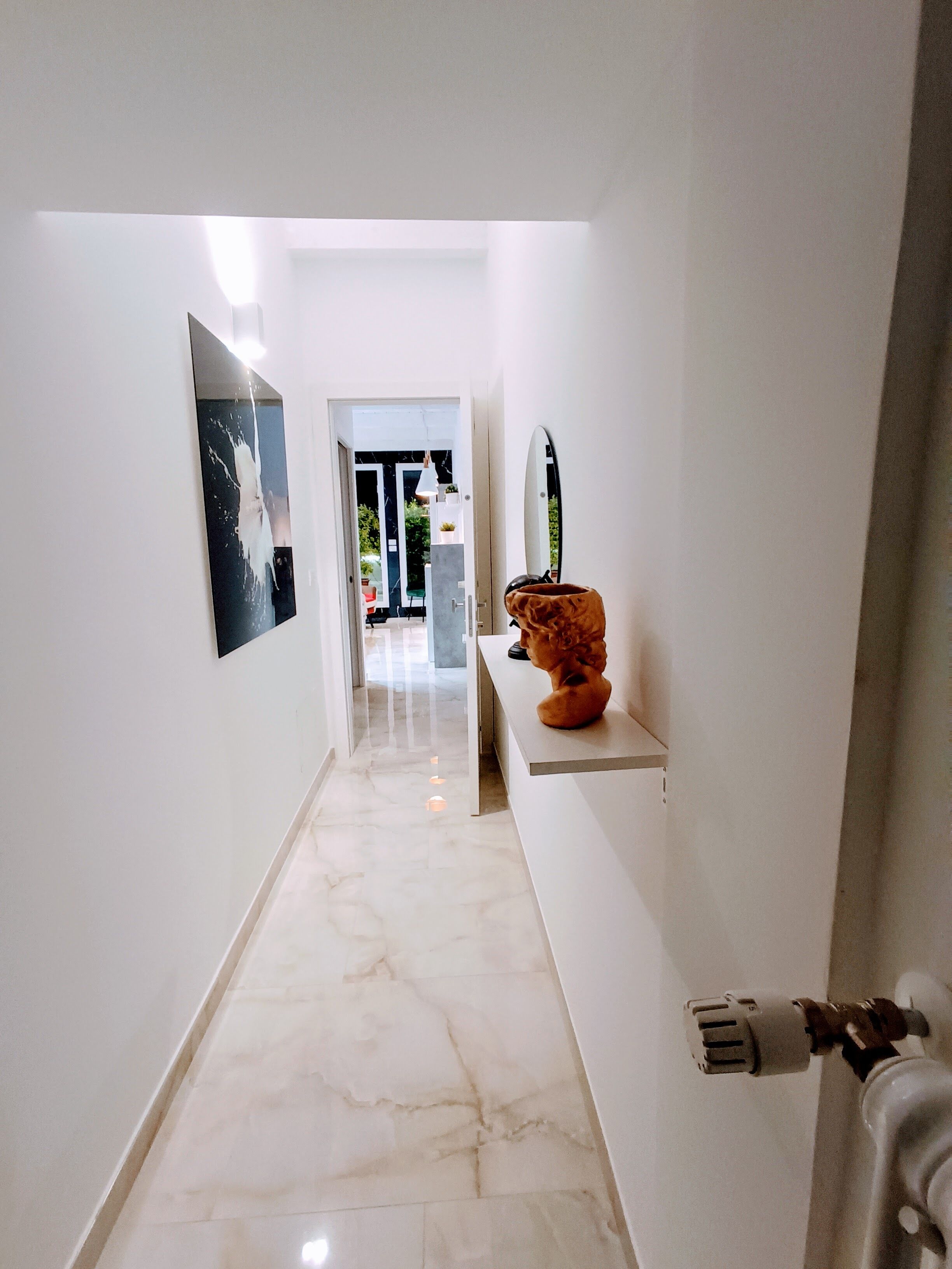 A hallway with a marble floor, a sculpture on a console table, and a door at the end leading to a room with a plant and a hanging light fixture.