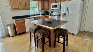A modern kitchen with wooden cabinets, a central island with stools, a stainless steel refrigerator, and a microwave. - Cottage on Snug Harbor, minutes from O.C. beaches, Berlin, and Assateague.  (Berlin)