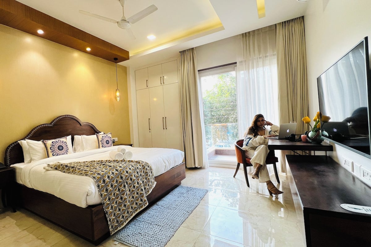 A modern hotel room with a large bed, a desk with a laptop, a flat-screen TV, and a balcony with curtains.