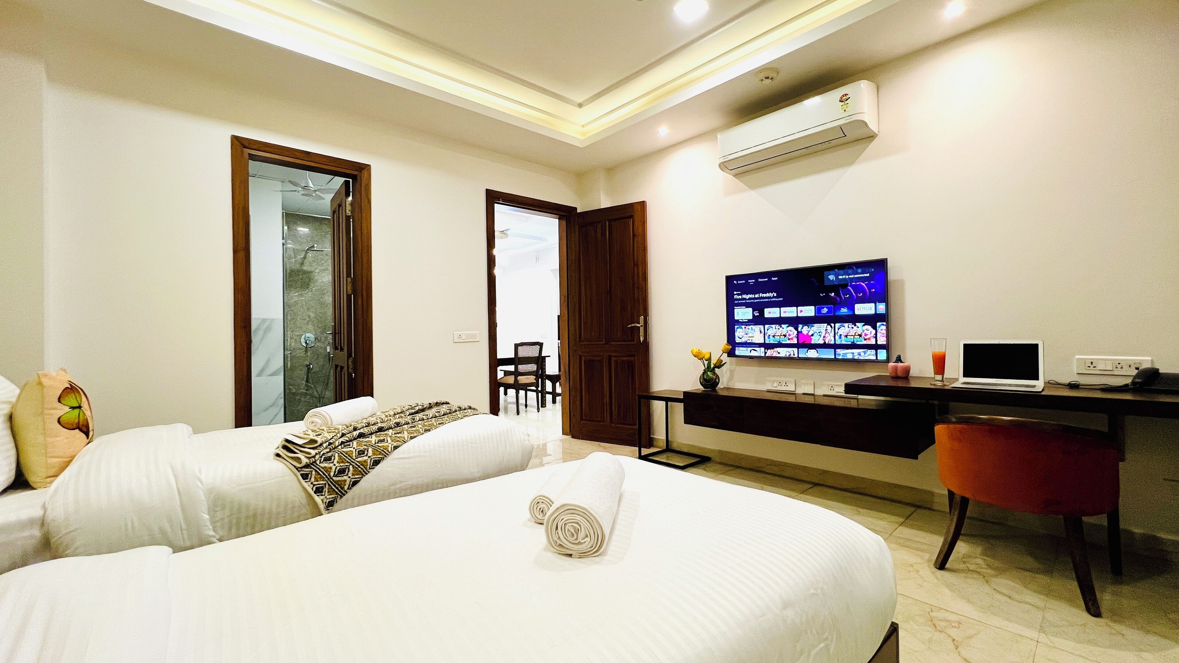 A hotel room with two beds, a desk with a laptop, a TV displaying a streaming service, and an air conditioning unit.
