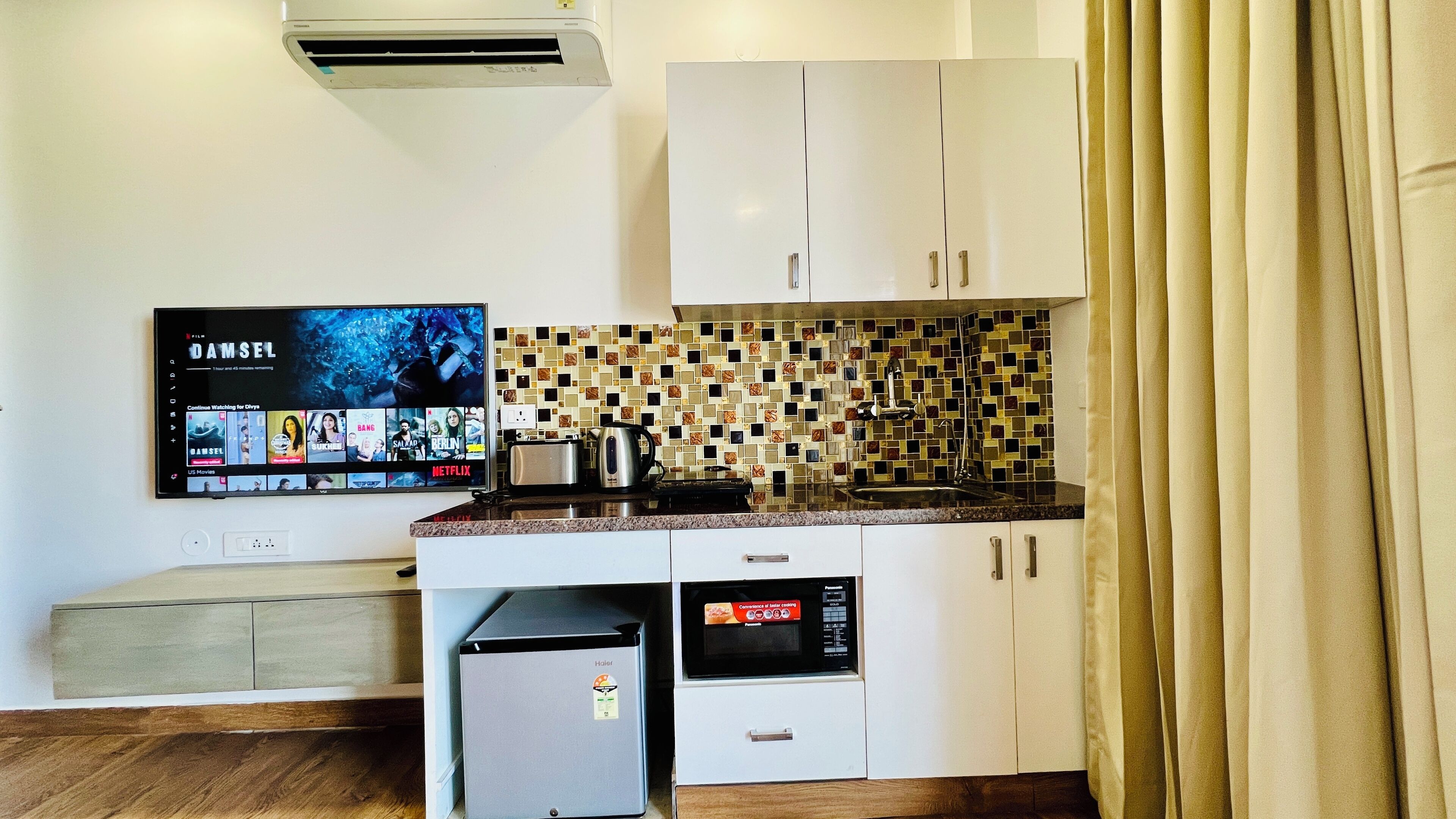 Cocina compacta con armarios blancos, salpicadero de azulejos, microondas, tostadora y un televisor de pantalla plana mostrando la interfaz de una plataforma de streaming.