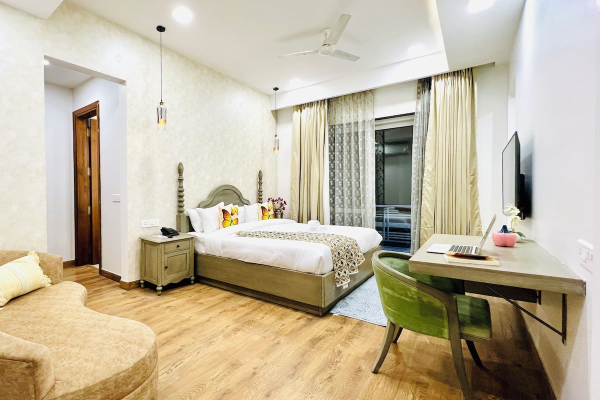 A hotel room with a bed, a desk with a laptop, a chair, a bedside table, a ceiling fan, and a window with curtains.