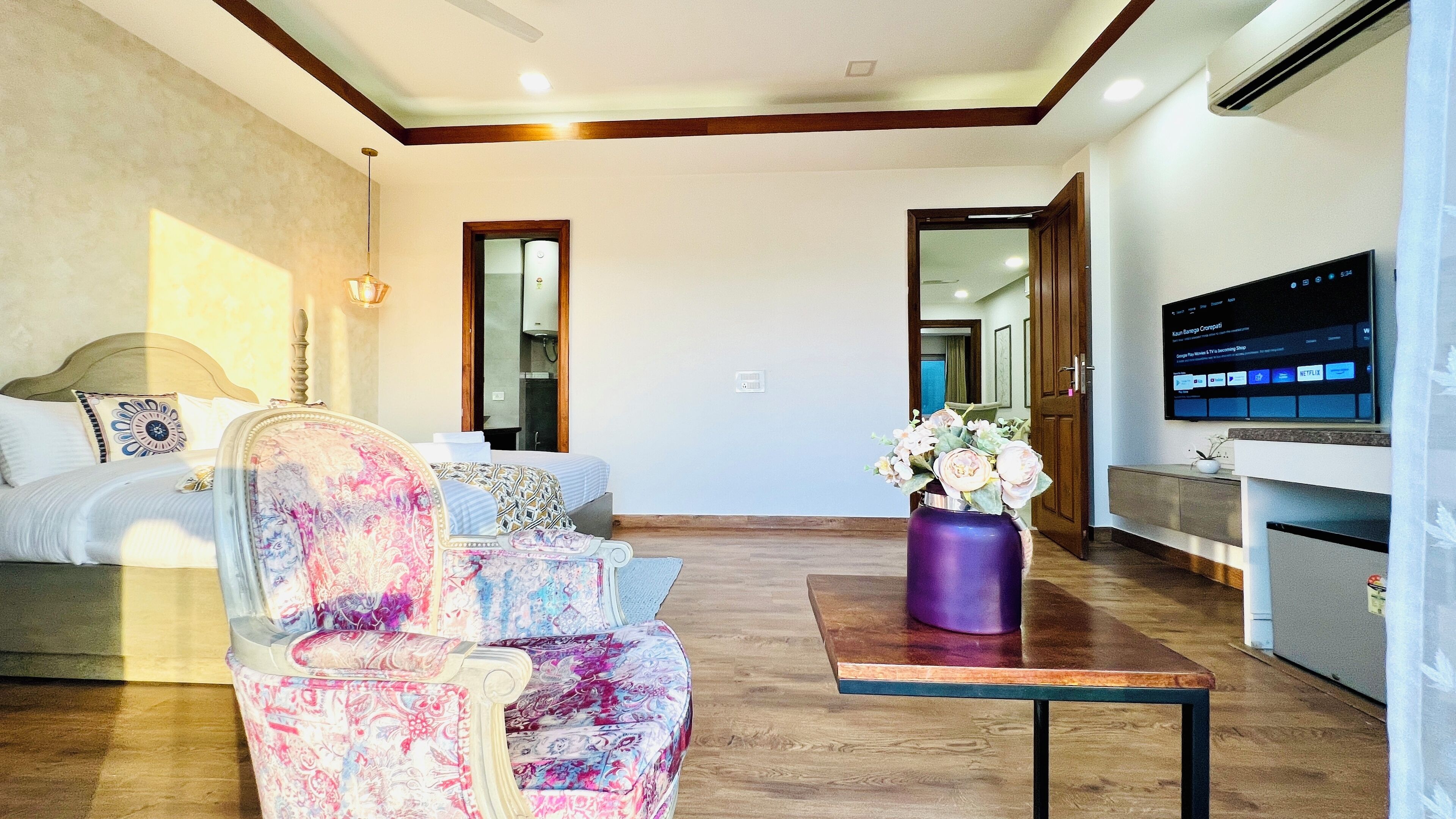 A hotel room with a bed, a sofa, a coffee table with a vase, a TV, and an air conditioning unit.