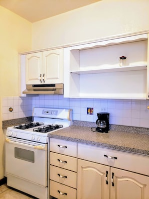 Een keuken met witte kastjes, een fornuis, een oven, een koffiezetapparaat en een betegelde achterwand.
