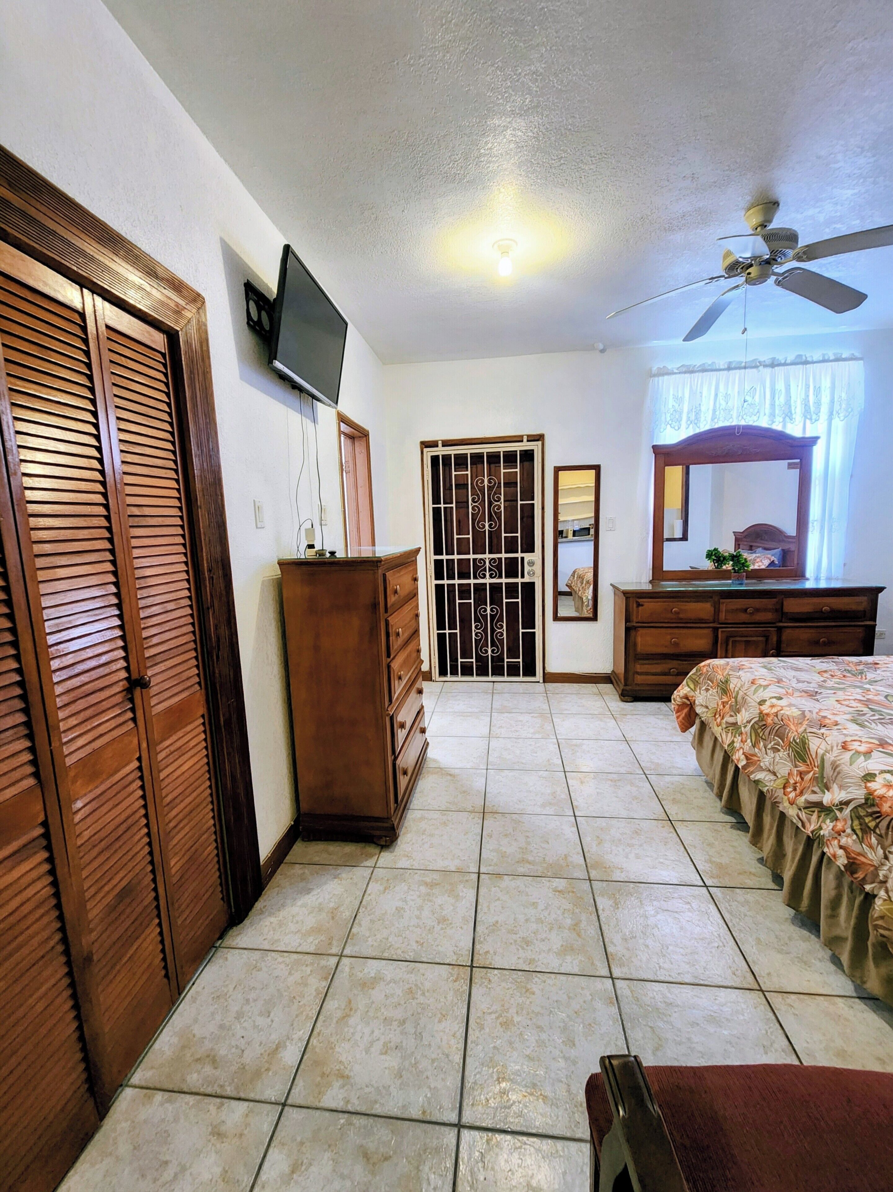 A bedroom with a bed, dresser, wardrobe, ceiling fan, TV, and a door with a decorative grille.