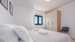 Een net opgemaakt bed met een witte deken en kussens, een raam met gordijnen, een houten kledingkast en een stoel.