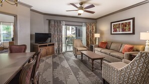A hotel room with a dining table, chairs, a sofa, a television, a ceiling fan, and a balcony with curtains.