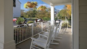 A porch with white chairs and a white railing, overlooking a street with parked cars and trees.