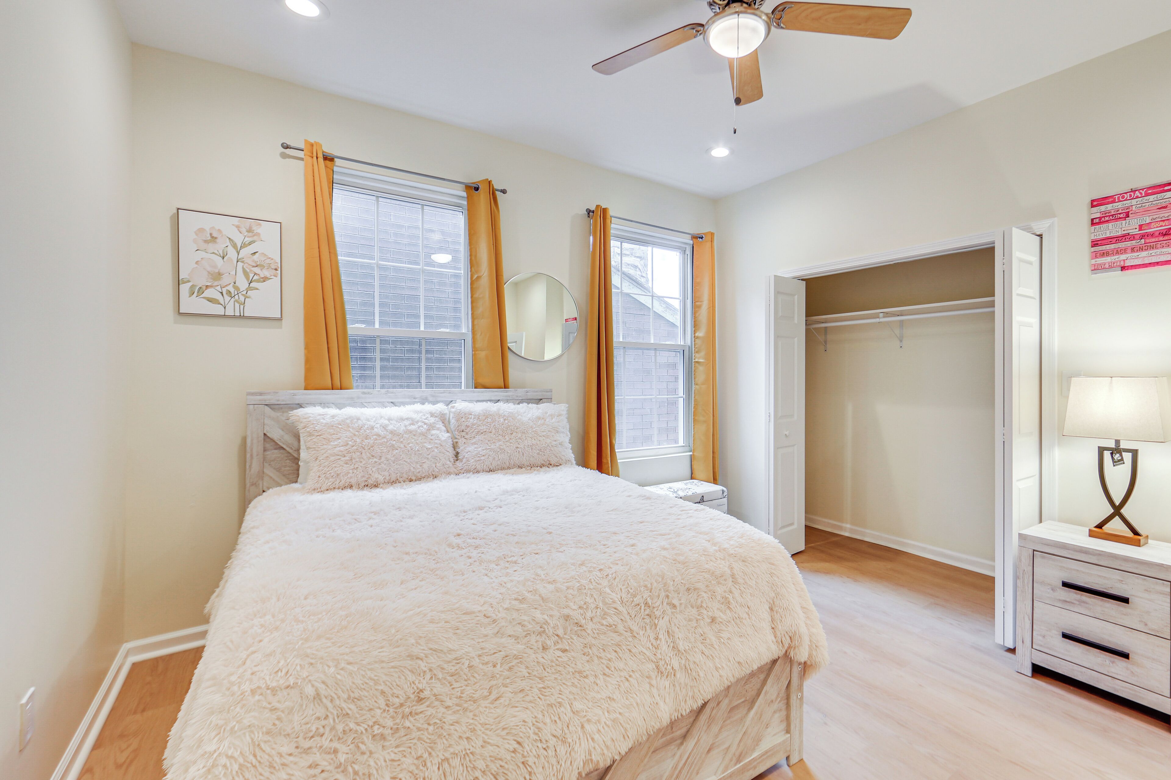 A bedroom with a bed, a nightstand, a ceiling fan, a window with curtains, a closet, and a wall-mounted mirror.