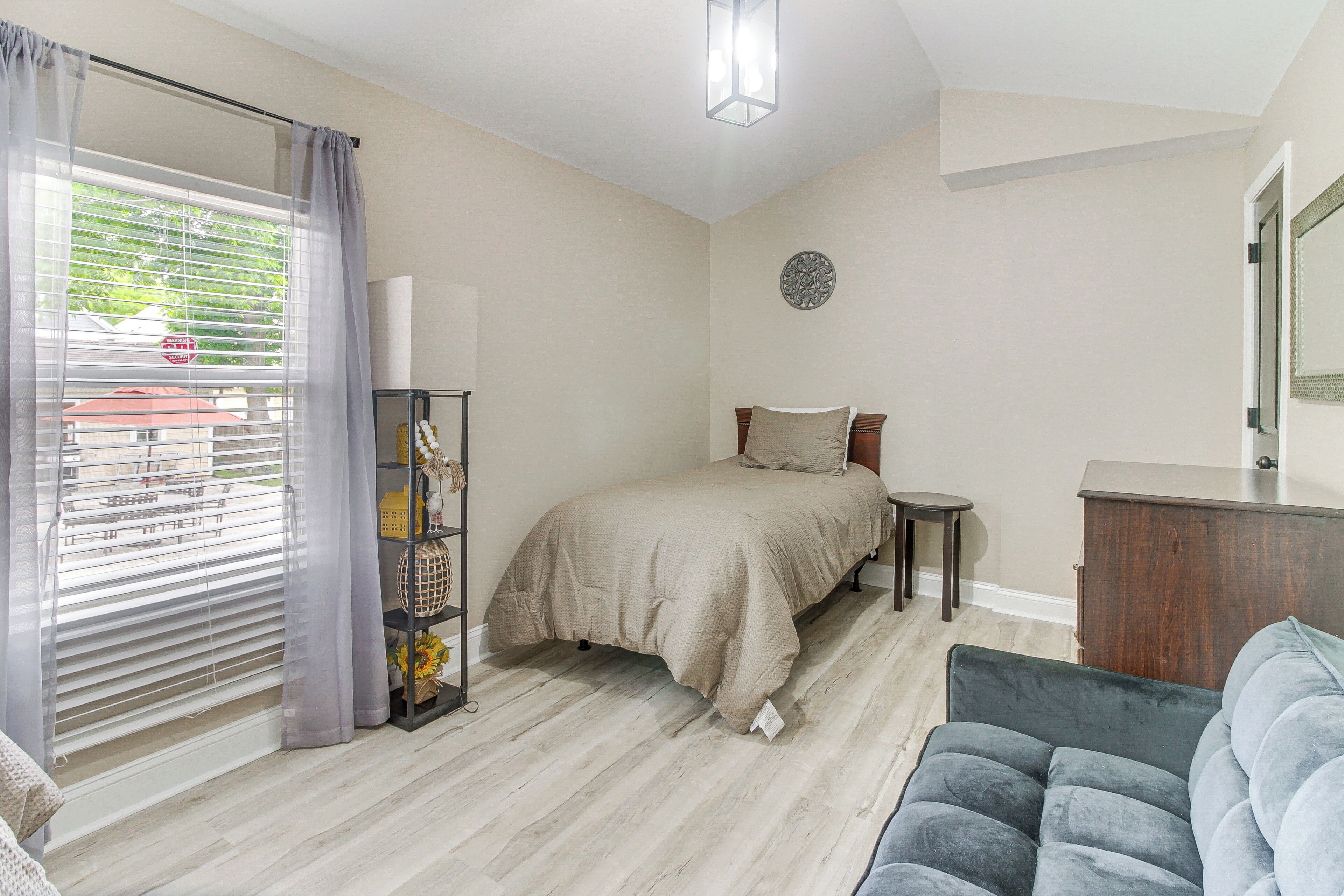 Ein Schlafzimmer mit Bett, Nachttisch, Kommode, Stuhl und einem Fenster mit Vorhängen.