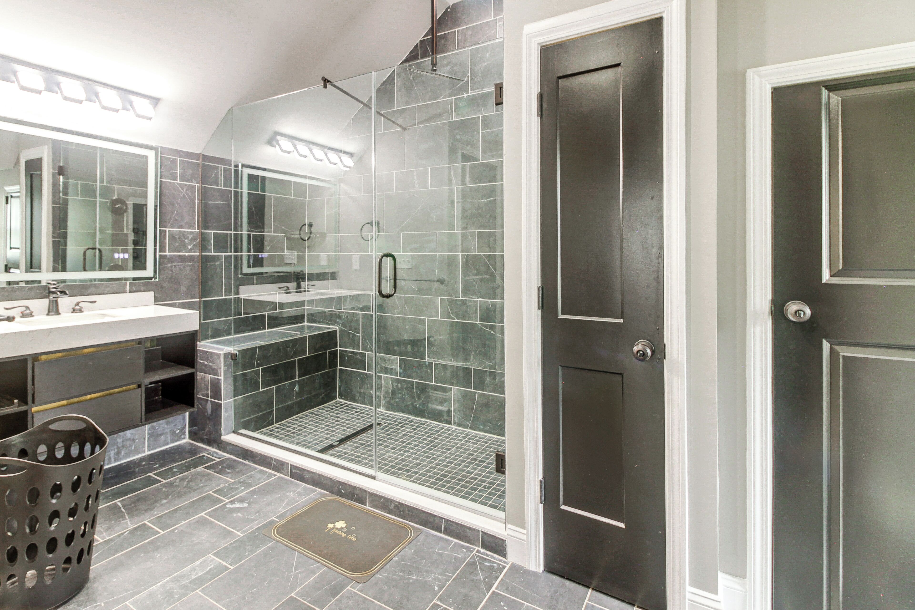 A modern bathroom with a glass-enclosed shower, a large mirror, and a dark-toned tiled floor.