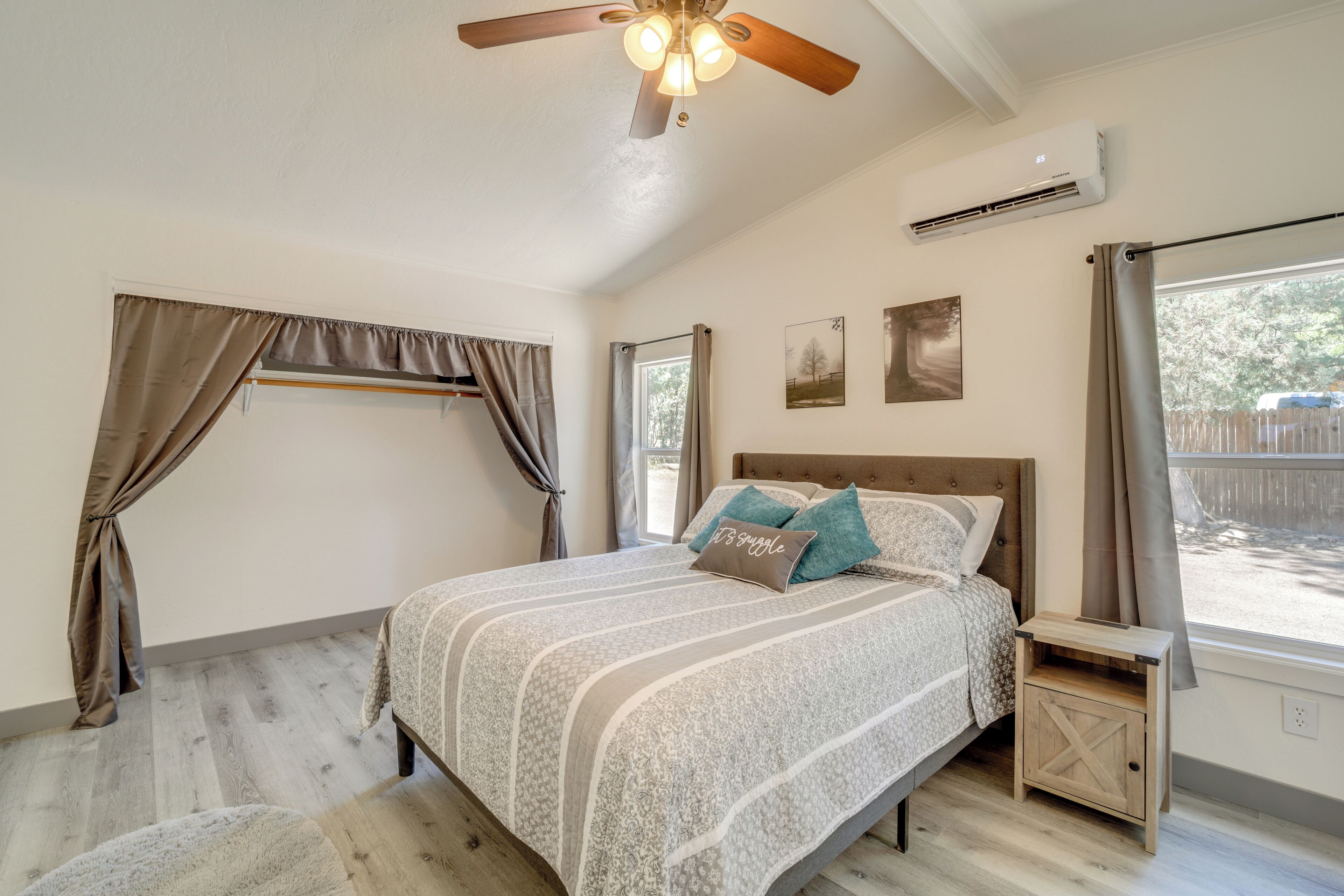 A bedroom with a bed, a ceiling fan, an air conditioning unit, a window with curtains, and a small wooden nightstand.