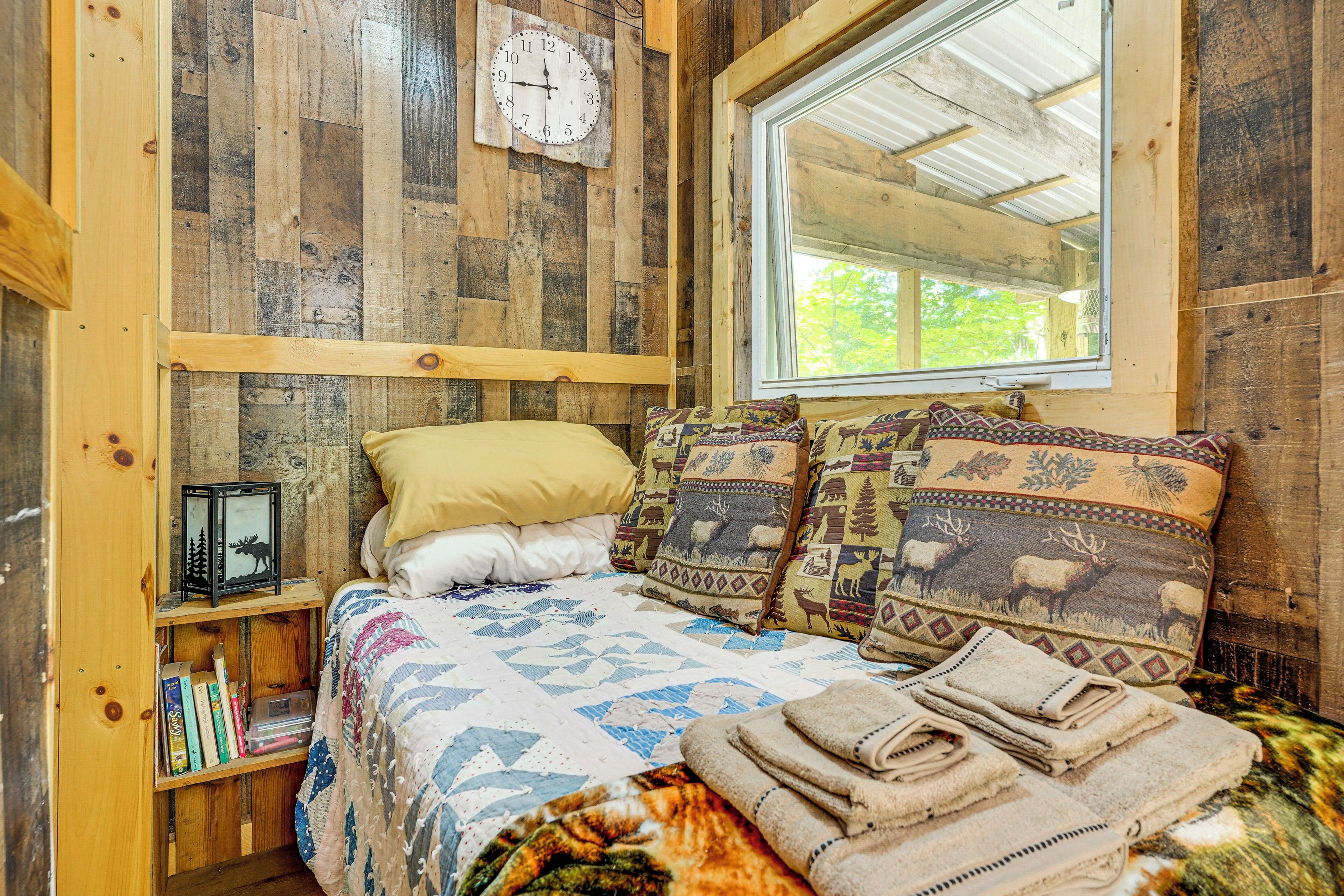 A rustic wooden room with a bed, a clock, a window, and a shelf with books.