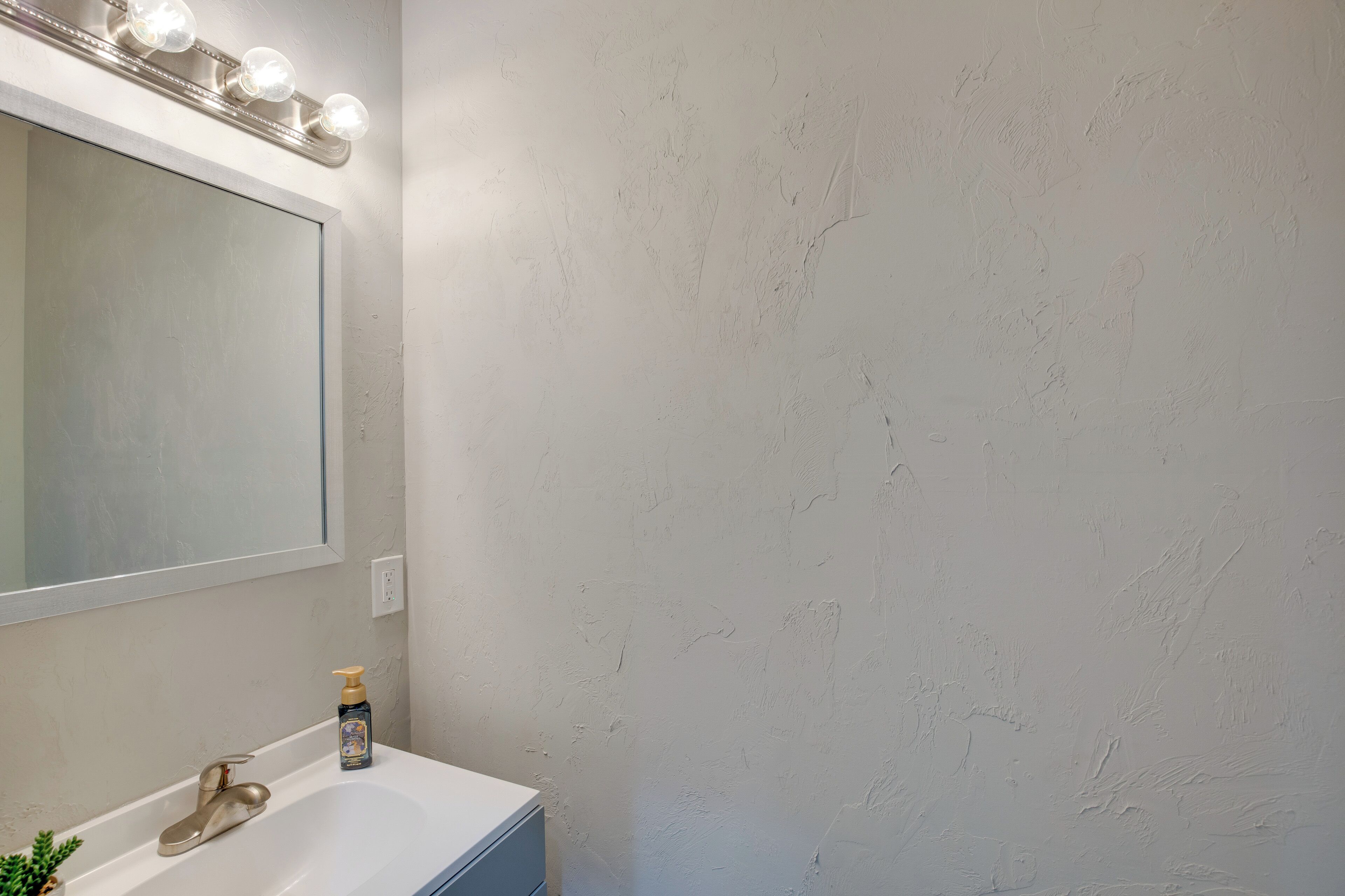 Une salle de bain avec un lavabo blanc, un miroir avec éclairage intégré et un mur à texture.