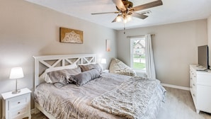 A bedroom with a bed, nightstands, a ceiling fan, a window with curtains, and a television on a dresser. - Walk to Lewisville Lake: Dog-friendly Texas Home (Little Elm)