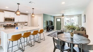 A modern kitchen with white cabinets, a central island with bar stools, a dining area with a round table, and a living room with a sofa and a plant. - Walk to Lewisville Lake: Dog-friendly Texas Home (Little Elm)