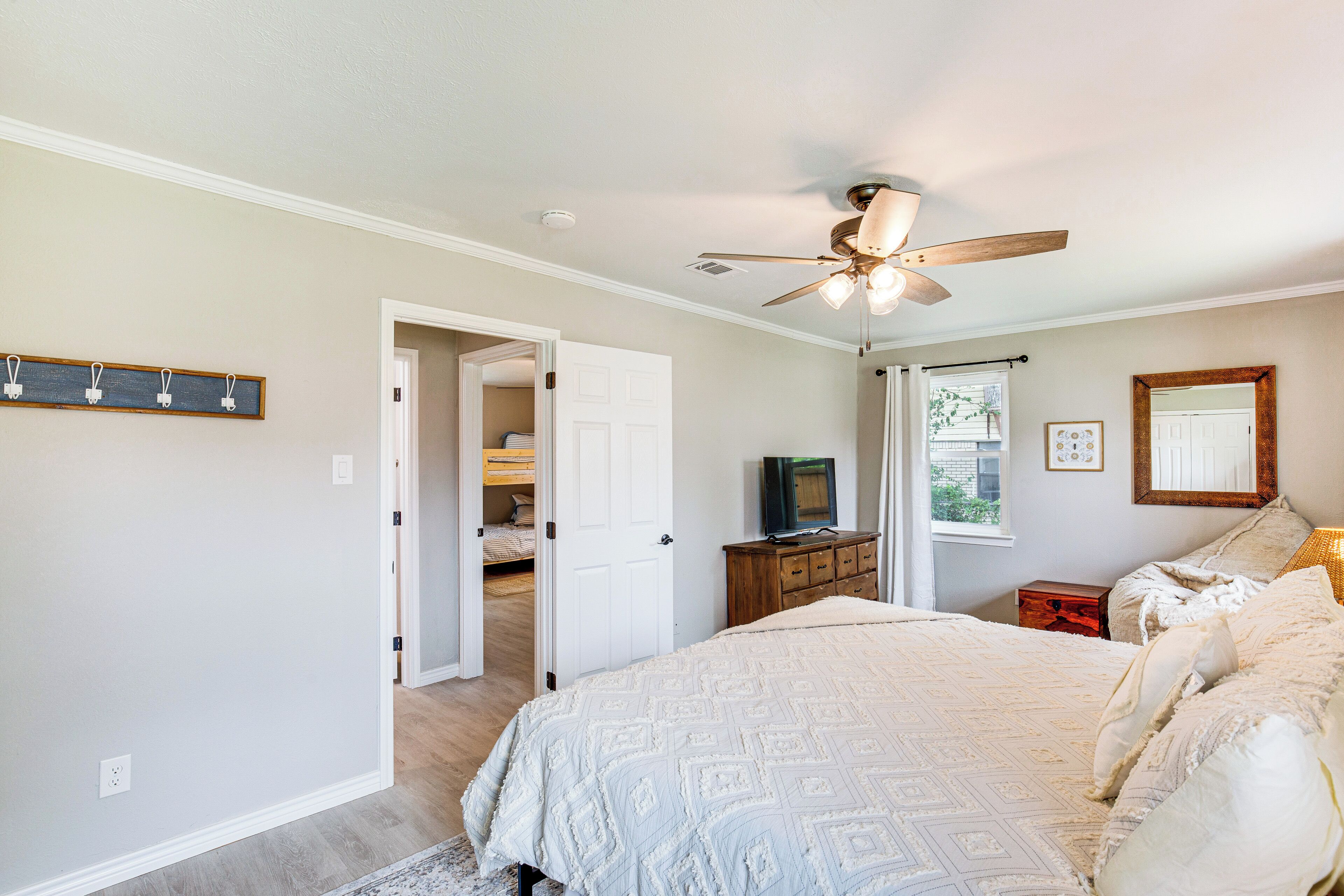 A bedroom with a large bed, a ceiling fan, a television on a wooden dresser, and a door leading to another room.