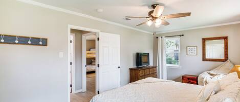 A bedroom with a large bed, a ceiling fan, a television on a wooden dresser, and a door leading to another room.