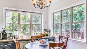 A dining area with a round table, wooden chairs, and a chandelier. Large windows provide views of greenery outside. - Lovely Mccormick Home in Savannah Lakes Village (McCormick)