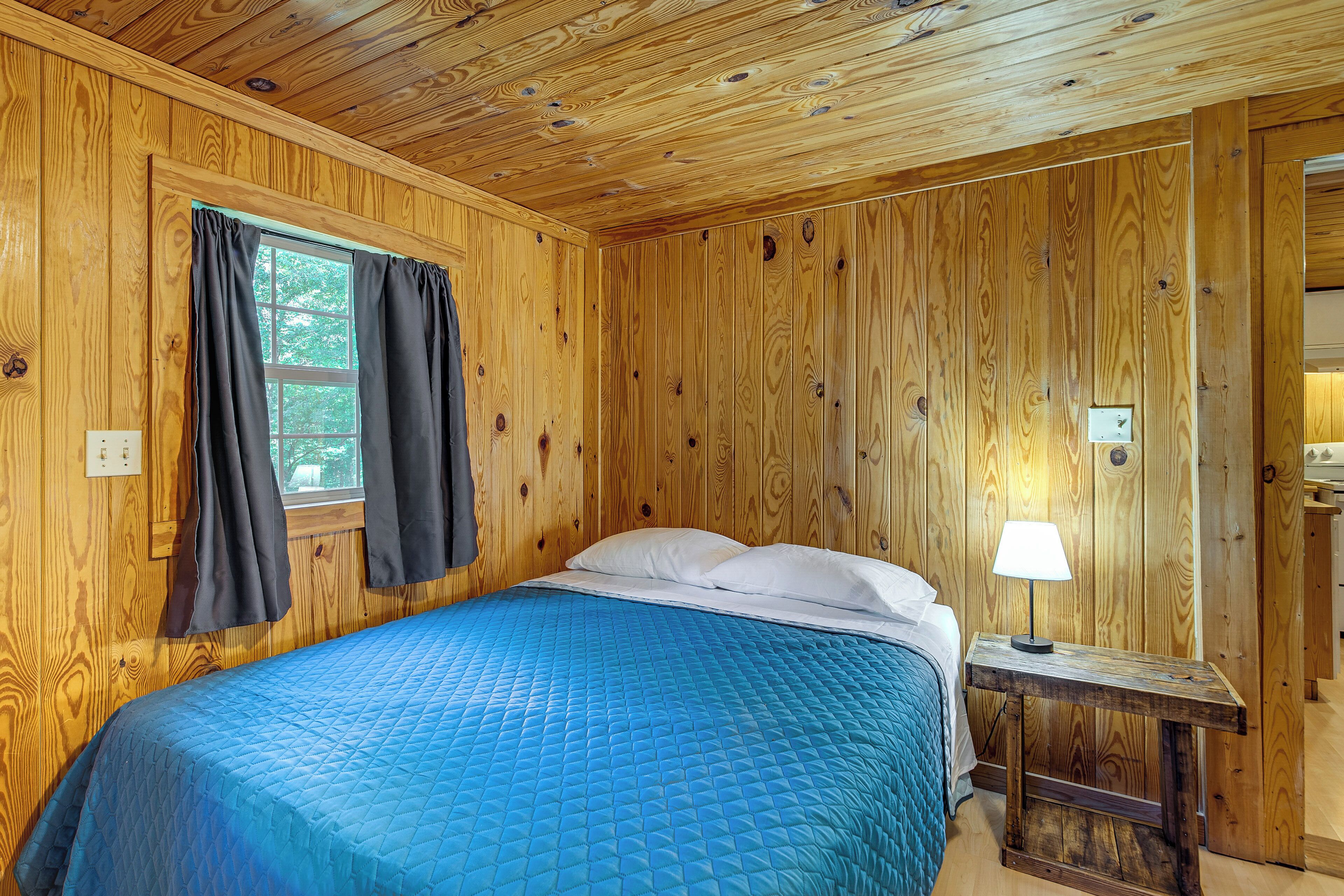 Una habitación con paneles de madera, una cama, un ventanal con cortinas y una mesita de noche con lámpara.