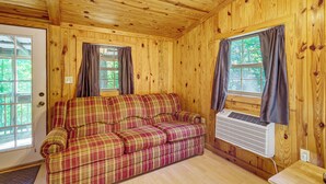 A wooden cabin interior with a plaid sofa, two windows with curtains, and an air conditioning unit. - Clarkson Cabin - 17 Mi to Mammoth Cave Natl Park! (Clarkson)