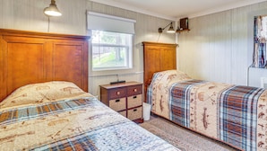 Un dormitorio con dos camas, cabeceros de madera, ventilador de techo, una ventana con persianas blancas y una mesita de noche con lámpara.