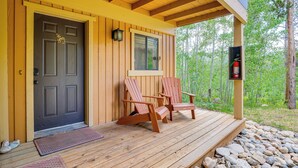 Une cabane en bois avec une porte d’entrée sombre, deux fauteuils Adirondack sur une terrasse en bois et un extincteur fixé au mur.