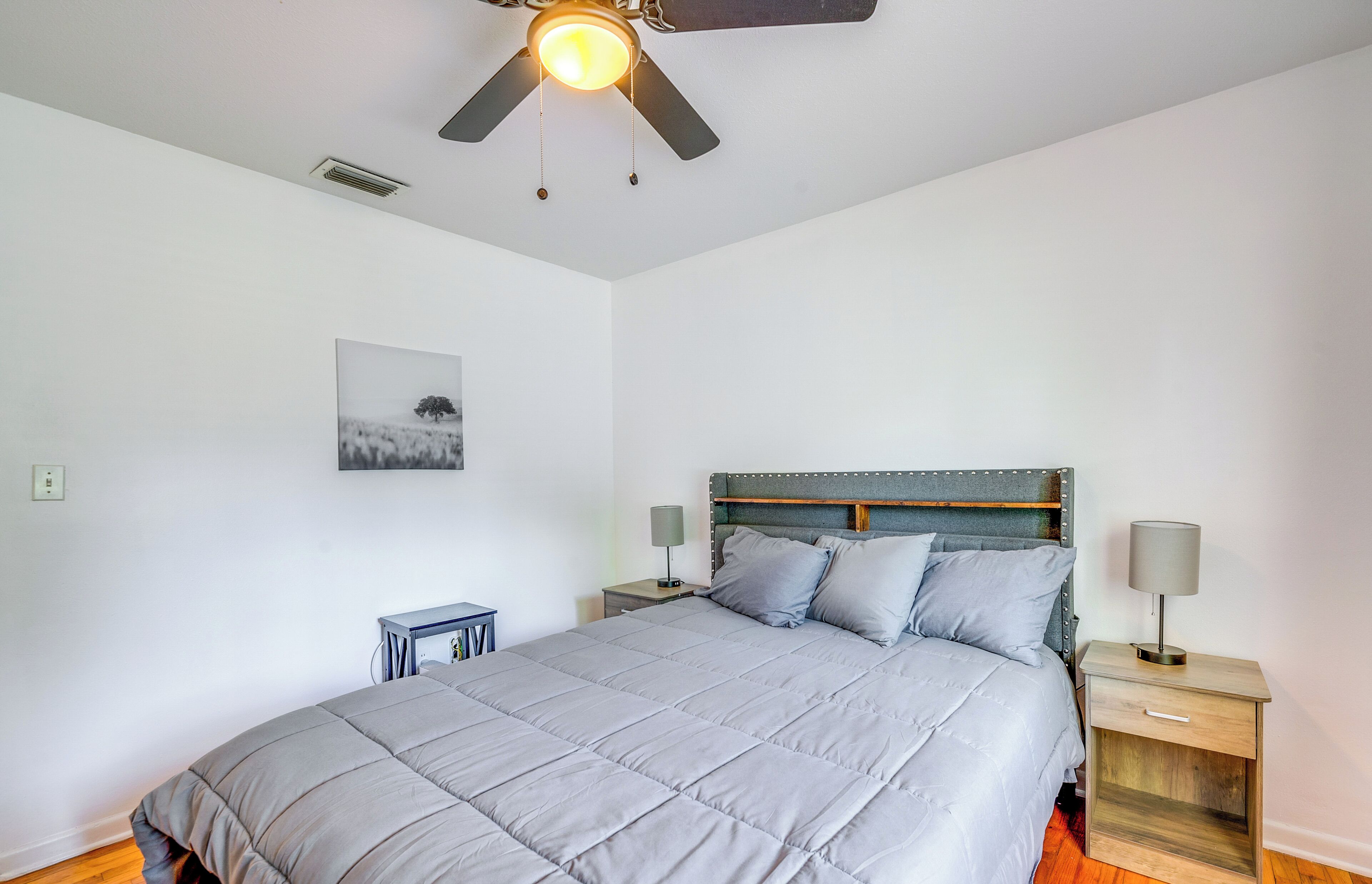 Een slaapkamer met een bed, een plafondventilator, een nachtkastje met een lamp en een aan de muur hangend schilderij.