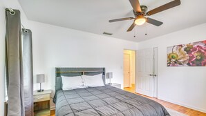 Une chambre à coucher avec un lit, un ventilateur de plafond, une table de chevet avec une lampe, une porte et un mur orné d’un tableau représentant des motifs floraux.