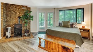 A bedroom with a brick fireplace, a bed with a green quilt, a wooden bench, and a large window with a forest view. - 'the Round House' - Treetop North Carolina Cabin! (Hendersonville)