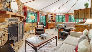 A cozy living room with a stone fireplace, wooden furniture, and large windows offering a view of the outdoors. - 'the Round House' - Treetop North Carolina Cabin! (Hendersonville)