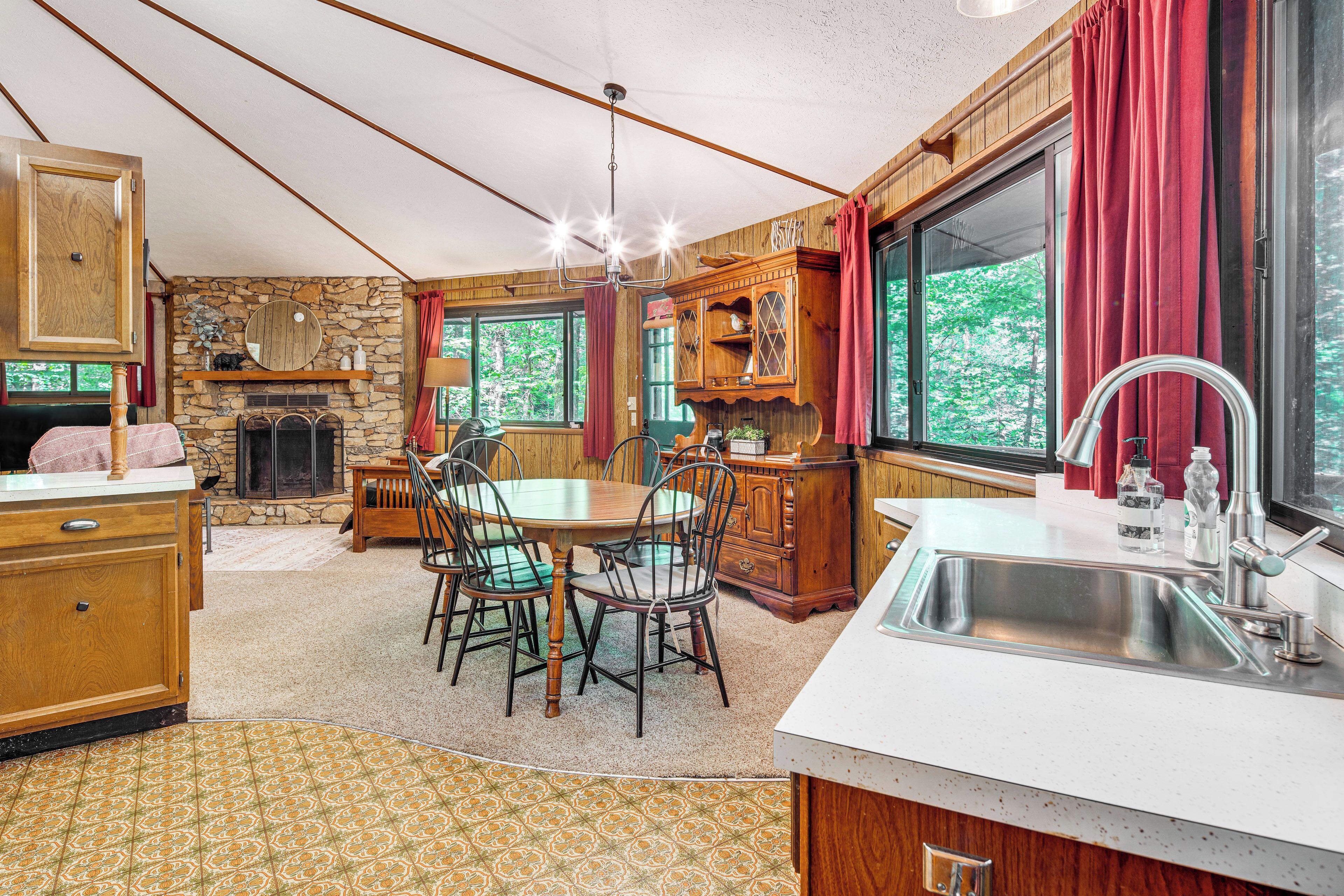 A cozy kitchen with a stone fireplace, wooden cabinets, and a large window with red curtains.