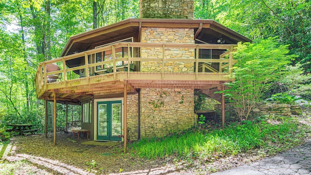 A rustic cabin with a stone chimney, wooden deck, and a door leading to the interior. - 'the Round House' - Treetop North Carolina Cabin! (Hendersonville)