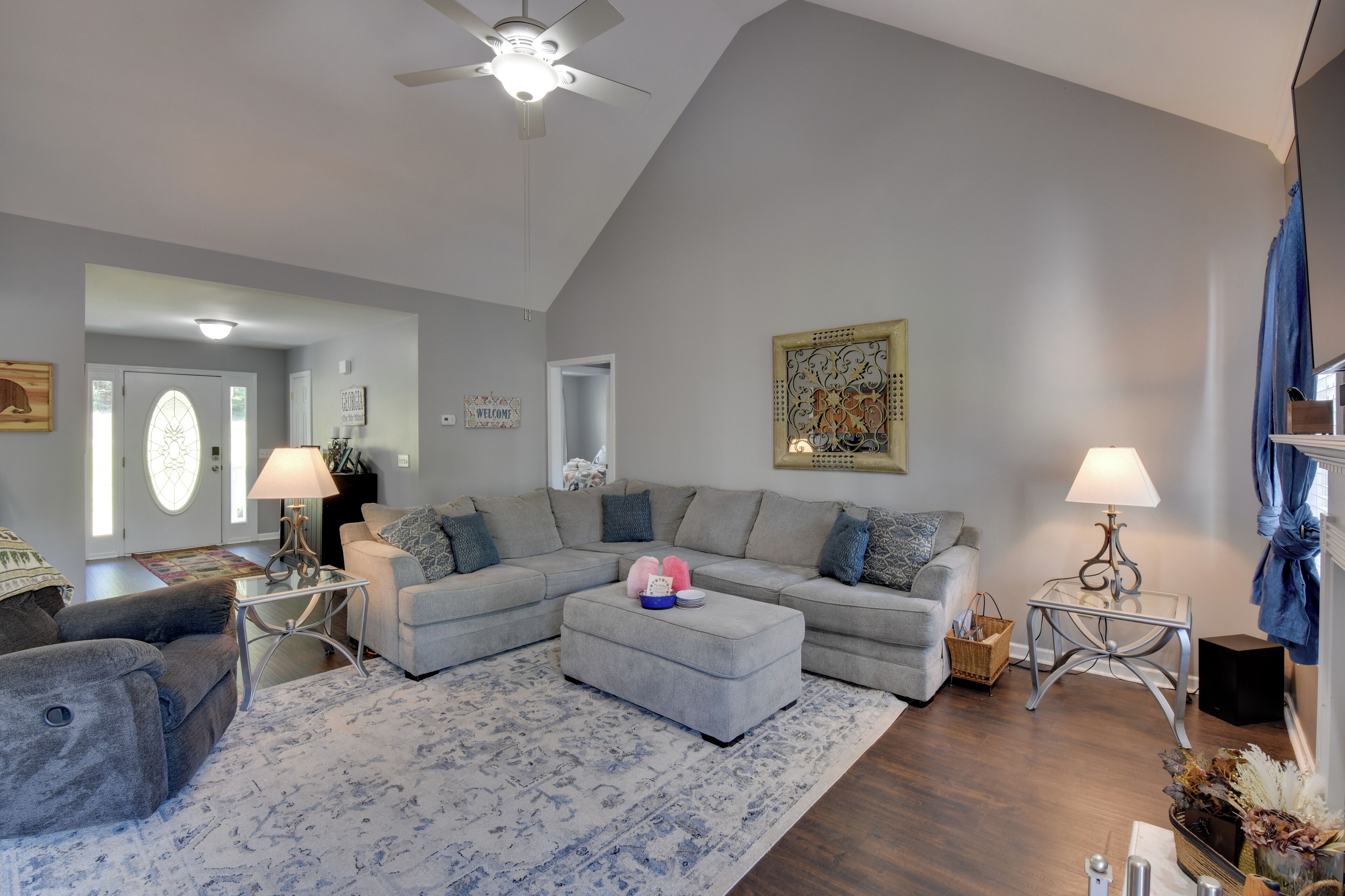 A living room with a grey sectional sofa, ottoman, and two side tables with lamps. There is a ceiling fan, a patterned rug, and a decorative wall piece.