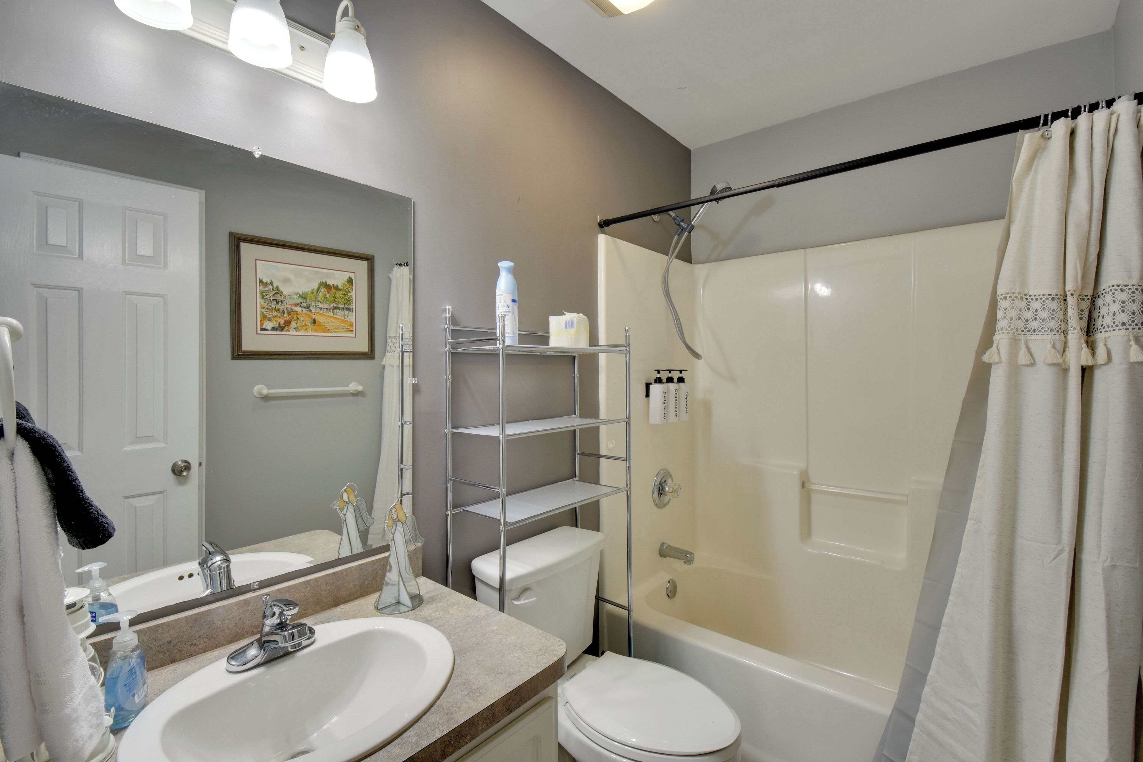 A bathroom with a double sink, toilet, and bathtub. There is a shelf with toiletries and a framed picture on the wall.
