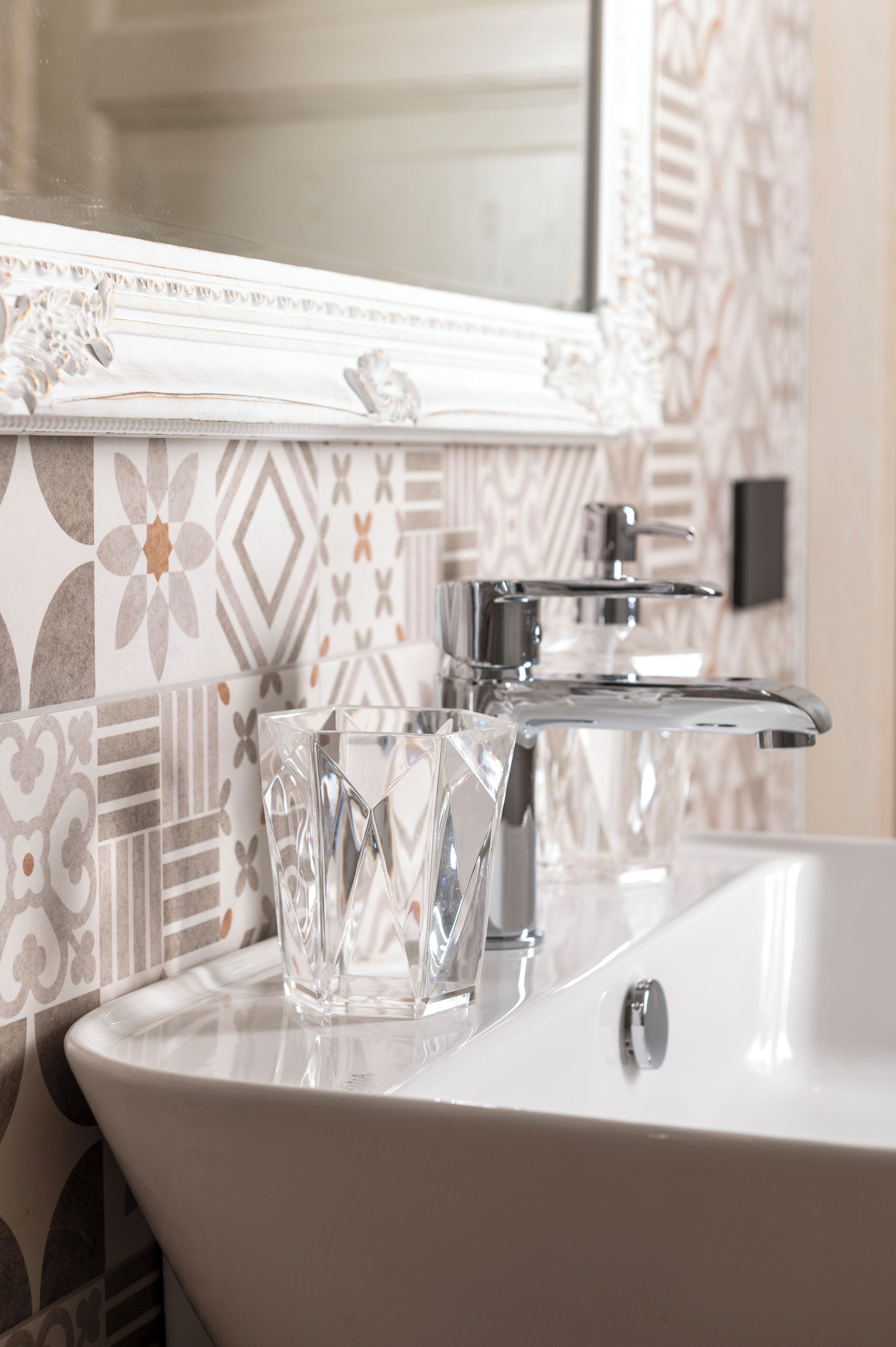 Une salle de bain avec un lavabo blanc, un vase en verre et des carreaux à motifs.