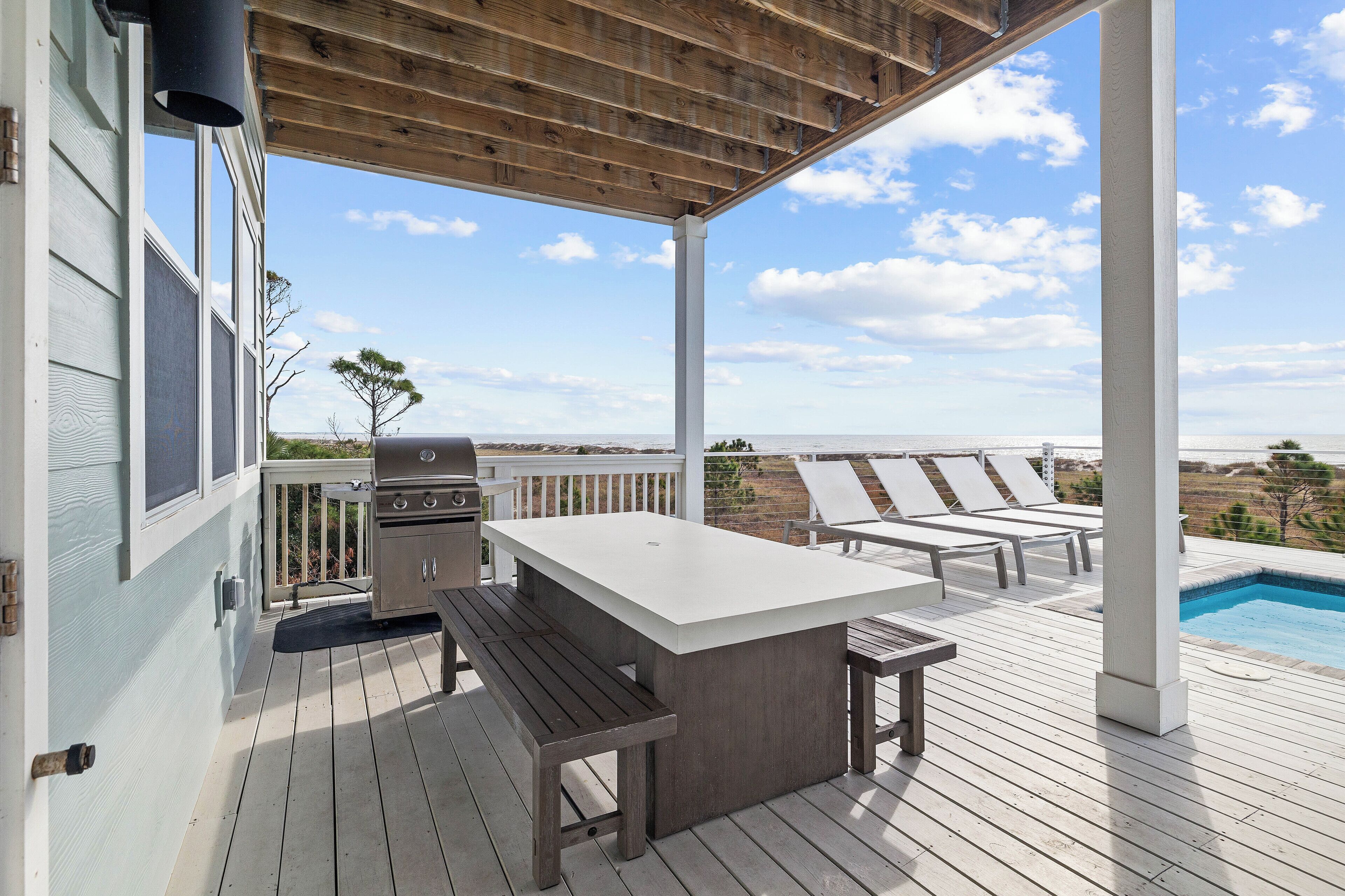A covered outdoor deck with a barbecue, a dining table, and lounge chairs overlooking a pool and ocean view.