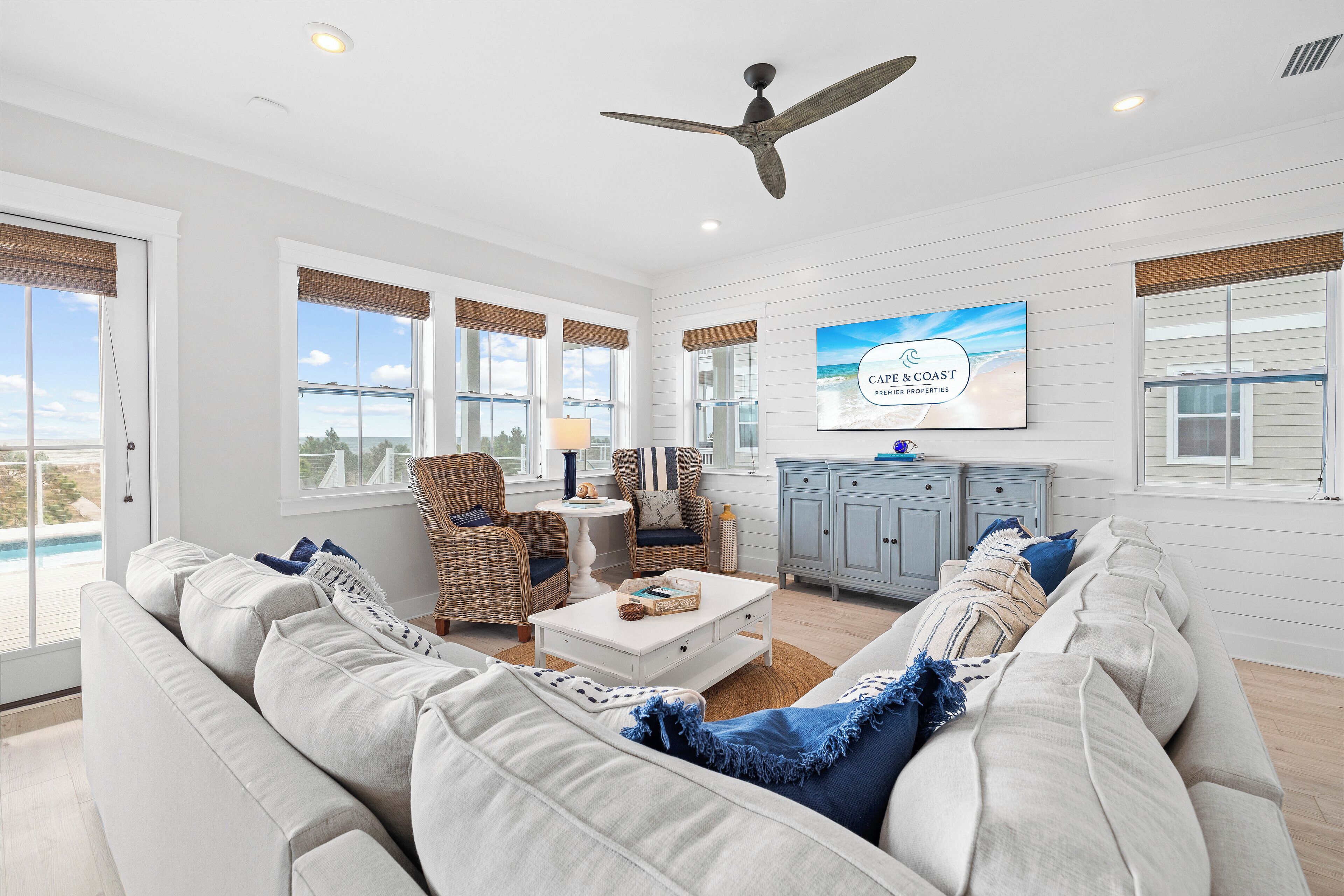 Ein großzügiger Wohnraum mit einem großen, grauen Ecksofa, einem weißen Couchtisch, Korbstühlen und einem Deckenventilator. Ein an der Wand befestigter Fernseher zeigt "Cape & Coast Properties".