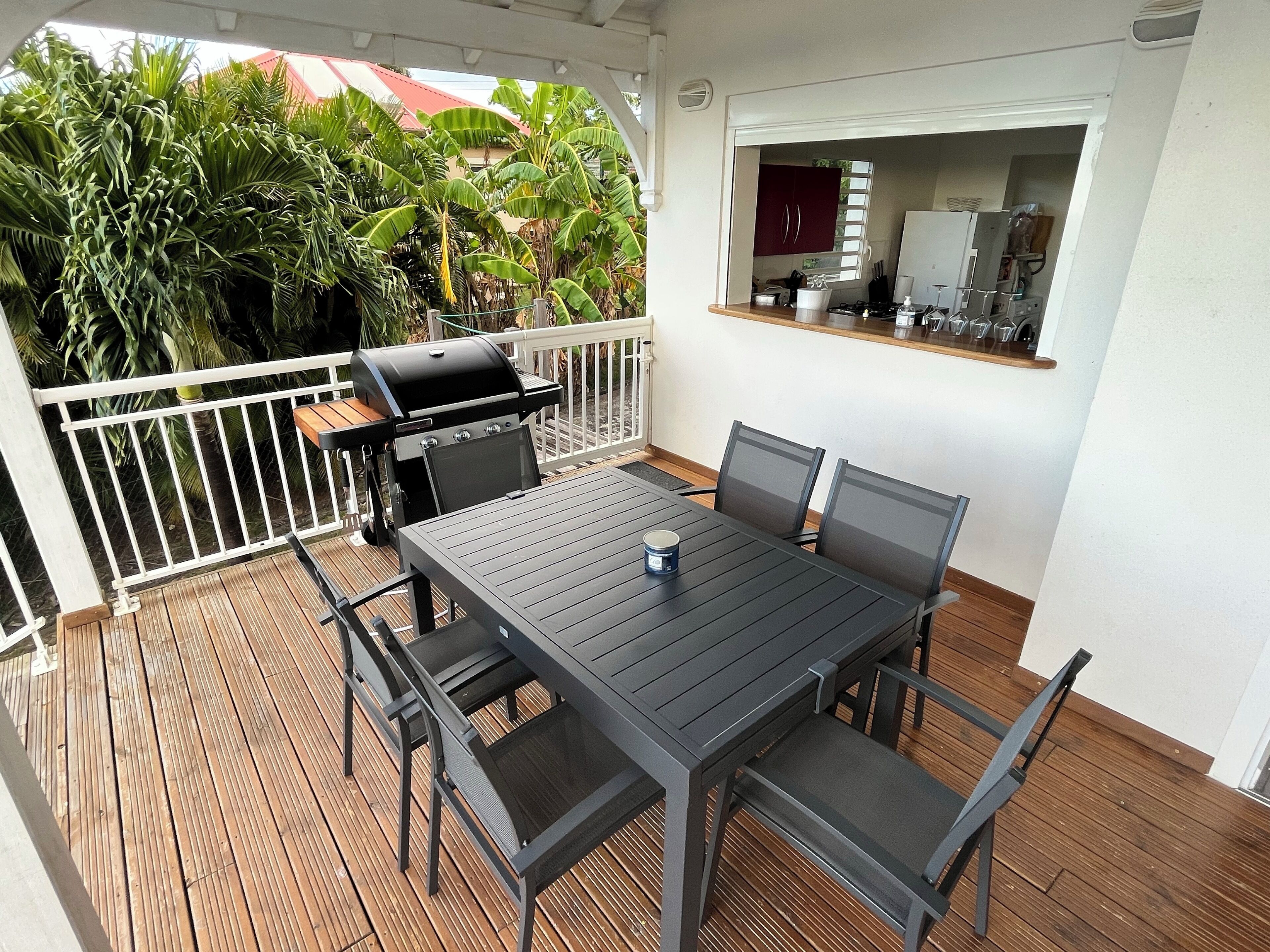A covered outdoor patio with a dining table and chairs, a barbecue grill, and a view of a house and trees in the background.