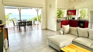 A spacious living area with a sofa, a wooden coffee table, and a dining set. A kitchen with red cabinets and a view of the ocean is visible through an open door. - Villa Tropical Quintet. 3 bedrooms. Sea view. Pool. Leroux beach at 800 m. (Deshaies)