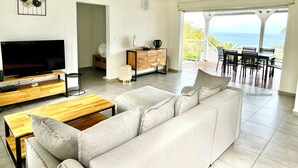A modern living room with a flat-screen TV, a wooden coffee table, a beige sofa, and a dining area with outdoor seating. - Villa Tropical Quintet. 3 bedrooms. Sea view. Pool. Leroux beach at 800 m. (Deshaies)